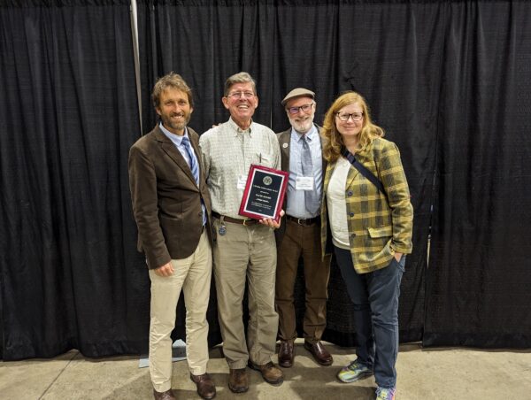 David Moore stands holding his Lifetime Achievement Award with colleagues surrounding him happily. 